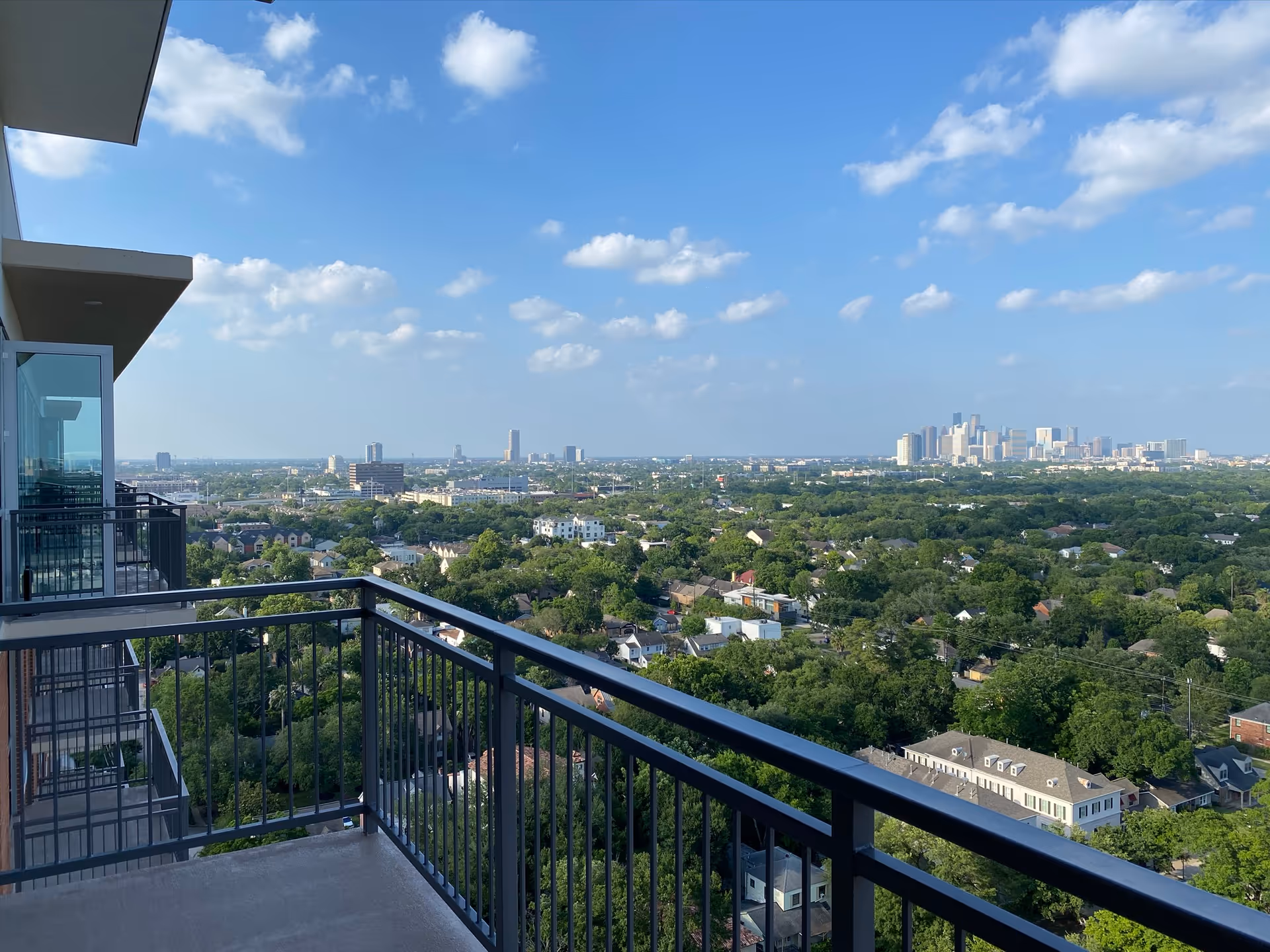 View from a high balcony overlooking a cityscape with many trees and buildings under a blue sky with scattered clouds.