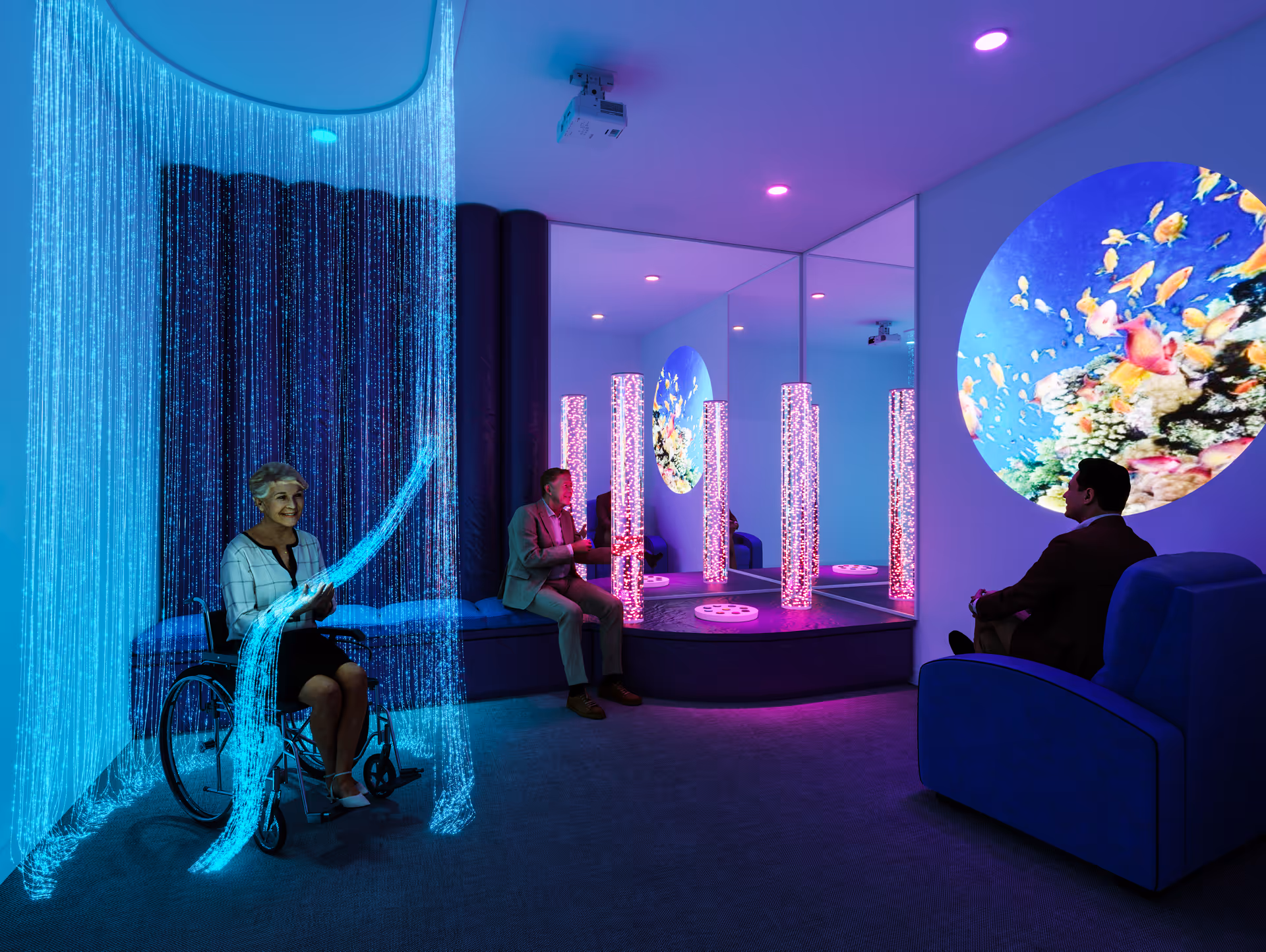 A sensory room with calming blue and purple lighting. An elderly woman in a wheelchair interacts with illuminated fiber optic strands, while two men sit on blue chairs and benches, observing a large circular screen displaying colorful fish and coral. The room features mirrored walls and vertical bubble tubes emitting pink light.