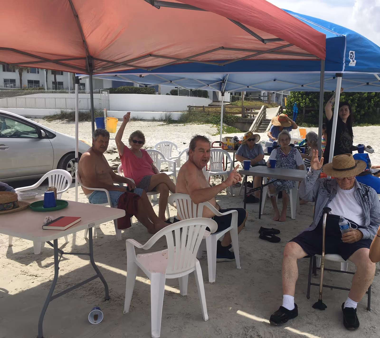 A group of elderly people sitting under red and blue canopy tents on a sandy beach. They are seated on white plastic chairs around folding tables, some holding drinks and raising their hands. A silver car is parked nearby, and buildings and greenery are visible in the background.