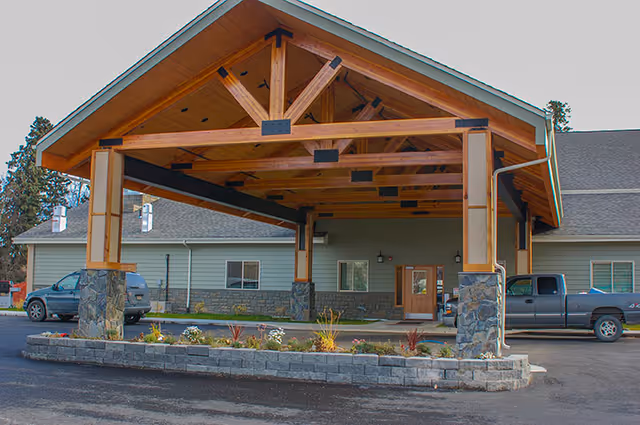 Entrance of a single-story assisted living facility with a large wooden covered porte-cochere supported by stone and wood pillars. There are vehicles parked on either side of the entrance and a small landscaped area with flowers and plants in front.