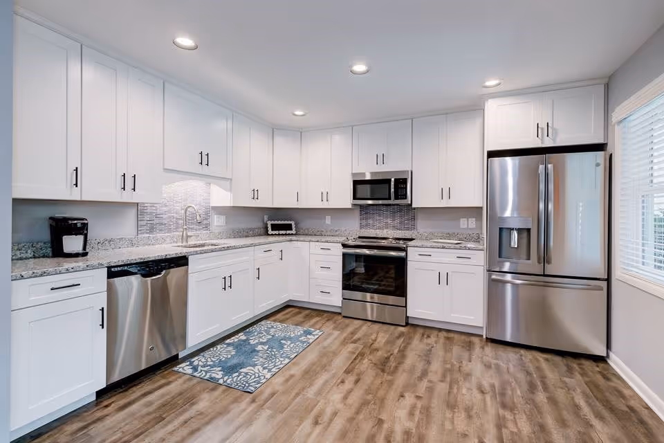Modern kitchen with white cabinets, stainless steel appliances including a refrigerator, oven, microwave, and dishwasher. The kitchen has granite countertops, a blue patterned rug on the wooden floor, recessed ceiling lights, and a window with blinds on the right side.