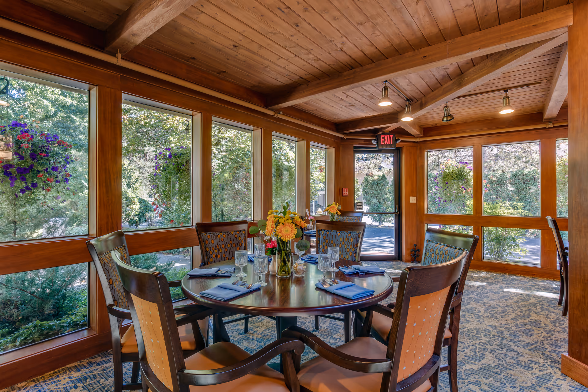Round dining table set with place settings and flowers in a sunlit room with wood-paneled ceiling and large windows overlooking a garden.