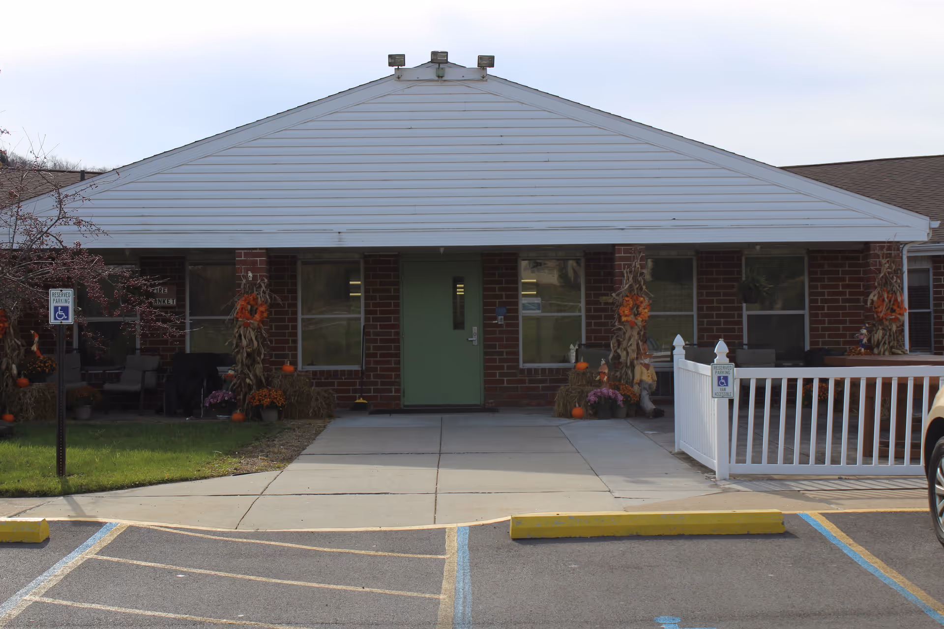 Front exterior view of a single-story brick building with a green door in the center, decorated with autumn-themed decorations including cornstalks, pumpkins, and flowers. There are two reserved handicap parking signs visible near the entrance and a white fence on the right side.