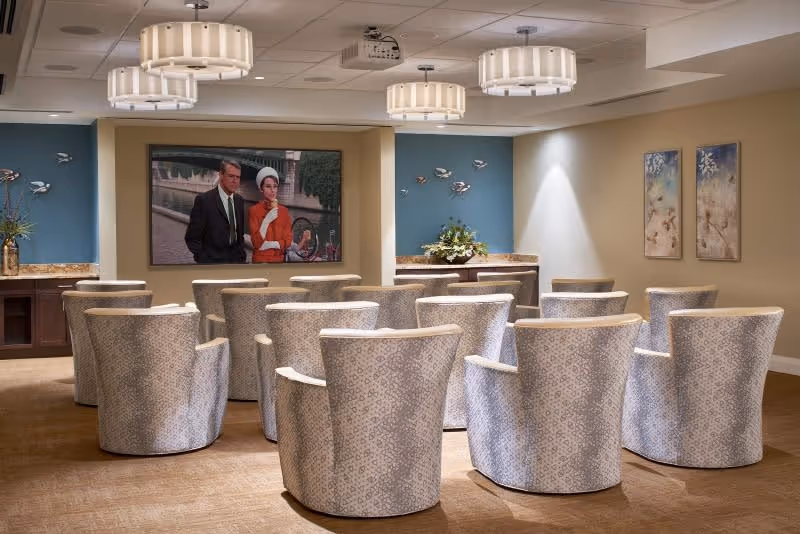 A cozy media room with multiple patterned armchairs arranged in rows facing a large wall-mounted television screen displaying a scene from a classic movie. The room features soft lighting from ceiling fixtures, decorative wall art, and a small counter with floral arrangements on either side of the TV.