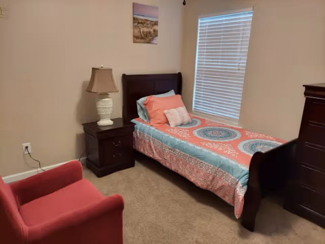 A small bedroom with a single bed featuring a colorful patterned bedspread and pillows. Next to the bed is a dark wooden nightstand with a white lamp. A red upholstered armchair is positioned in the corner. The room has beige walls, a window with white blinds, a framed picture on the wall, and a dark wooden dresser.