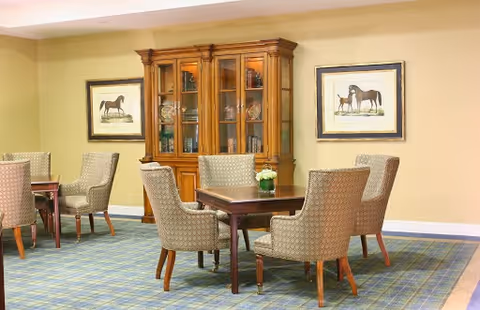 A cozy interior room with a wooden display cabinet filled with glassware and dishes, surrounded by framed horse artwork on the walls. There are several upholstered chairs arranged around wooden tables on a patterned carpeted floor.