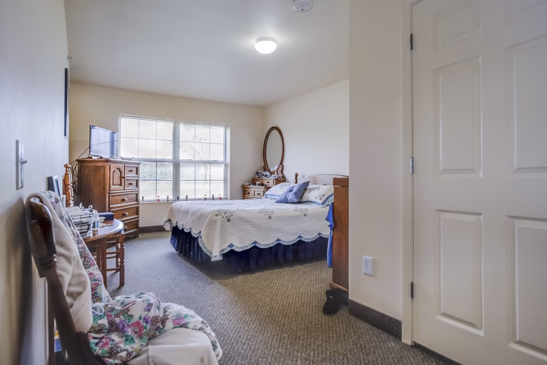 A cozy bedroom in Devonshire Retirement Village featuring a bed with a white and blue quilt, a wooden dresser with a round mirror, a wooden chest of drawers with a TV on top, a chair with floral cushions, and a large window letting in natural light.