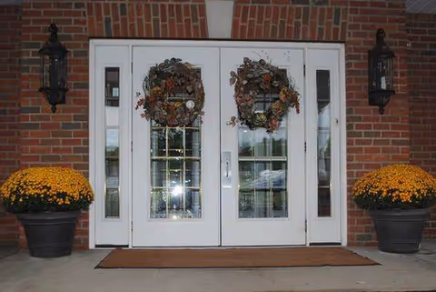 White double doors with wreaths flank a brick entrance and orange mums in pots.