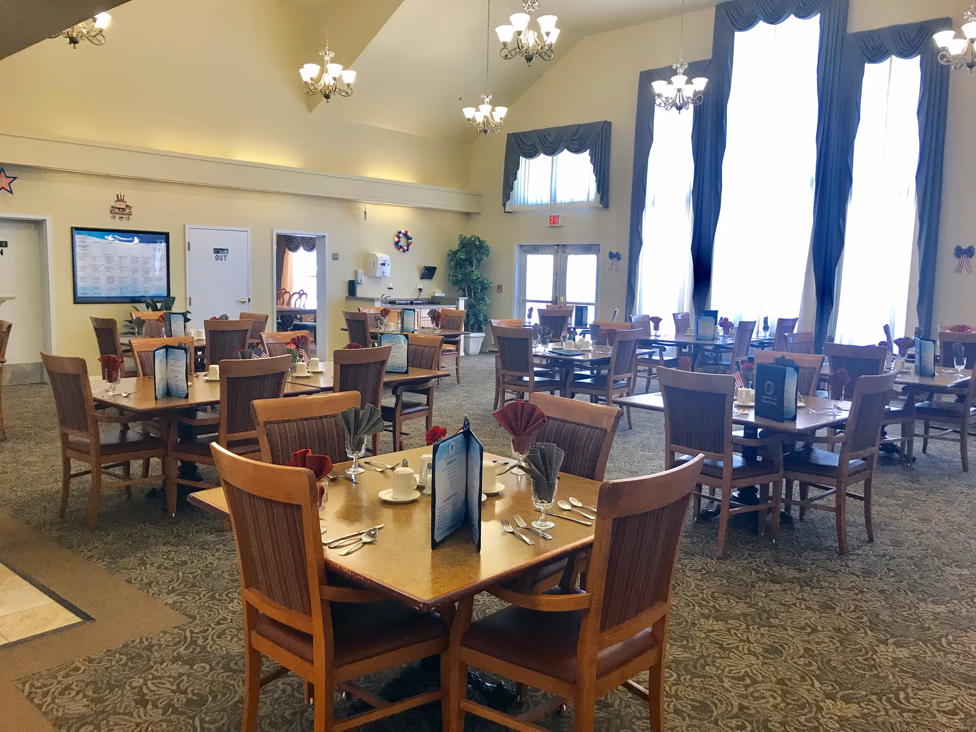 Spacious dining room with multiple set wooden tables and chairs, large windows with drapes, and chandeliers.