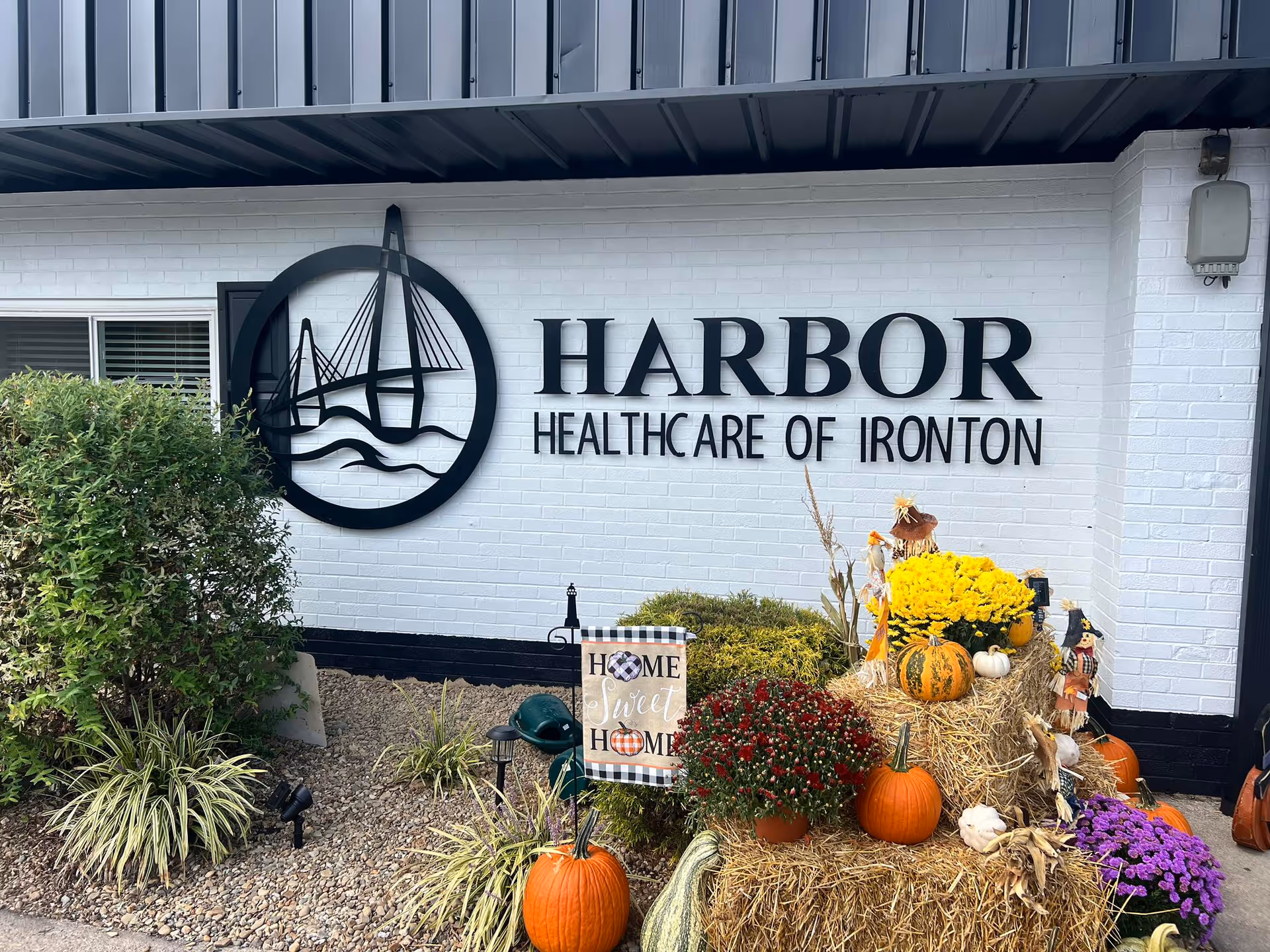 Exterior wall of Harbor Healthcare of Ironton with a large black metal sign featuring a bridge design and the facility name. In front of the wall, there are autumn decorations including pumpkins, hay bales, colorful flowers, and a small sign that reads 'Home Sweet Home'. There are also some green bushes and plants around the area.