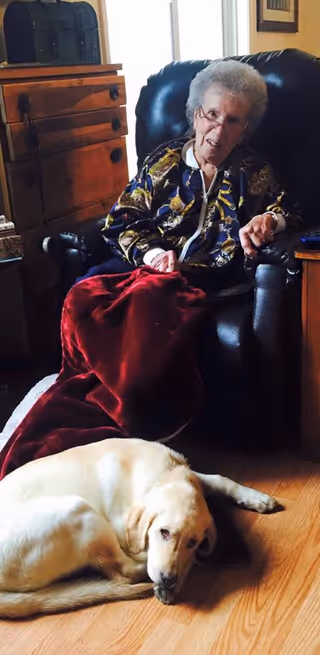 An older adult sits in a recliner with a red blanket while a yellow Labrador lies on the hardwood floor.