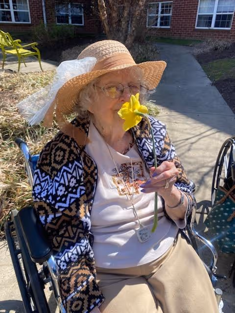 An elderly woman sitting in a wheelchair outdoors on a sunny day, wearing a wide-brimmed straw hat, glasses, a patterned cardigan, and beige pants, holding and smelling a yellow flower with a concrete walkway and brick building in the background.