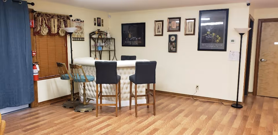 A cozy interior room with a small bar area featuring a white tufted counter and four bar stools, two with blue cushions and two with black cushions. The room has wooden flooring, cream-colored walls adorned with framed artwork, a tall floor lamp, a window with wooden blinds and decorative valance, and a metal shelf holding glassware and bottles. A fire extinguisher is mounted near the window, and there is a closed wooden door on the right side.