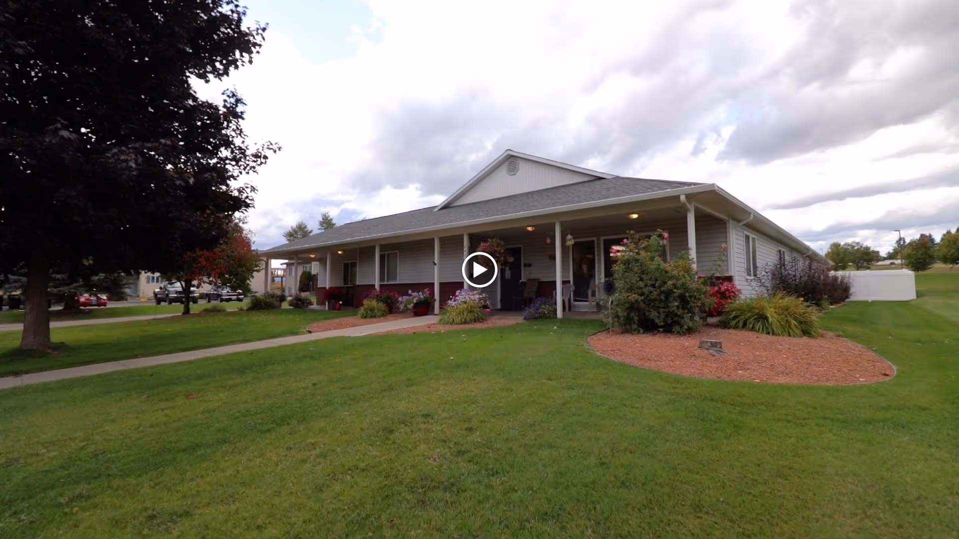 Single-story senior care facility building with a covered porch, surrounded by a well-maintained lawn and landscaped garden beds, under a cloudy sky.