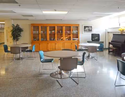 A senior living facility dining area with round tables, chairs, a wooden cabinet, and a piano.