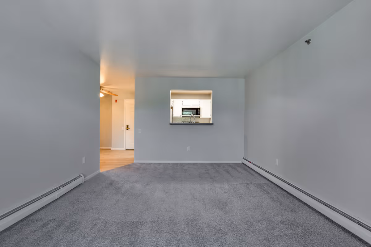 Empty room with gray carpet and white walls, featuring a small pass-through window to a kitchen with white cabinets and a microwave. The room has baseboard heating along the walls and a view of a hallway with a ceiling fan and a door in the background.