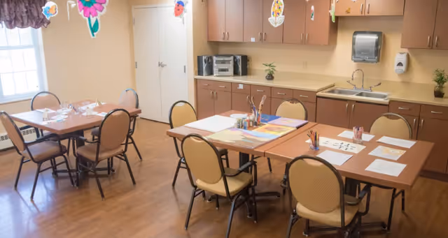 A well-lit room with two tables and several chairs arranged around them. The tables have papers, coloring supplies, and art materials on them. The room has wooden flooring, beige walls, and cabinets with a sink and a stereo system along one wall. Colorful paper decorations hang from the ceiling.