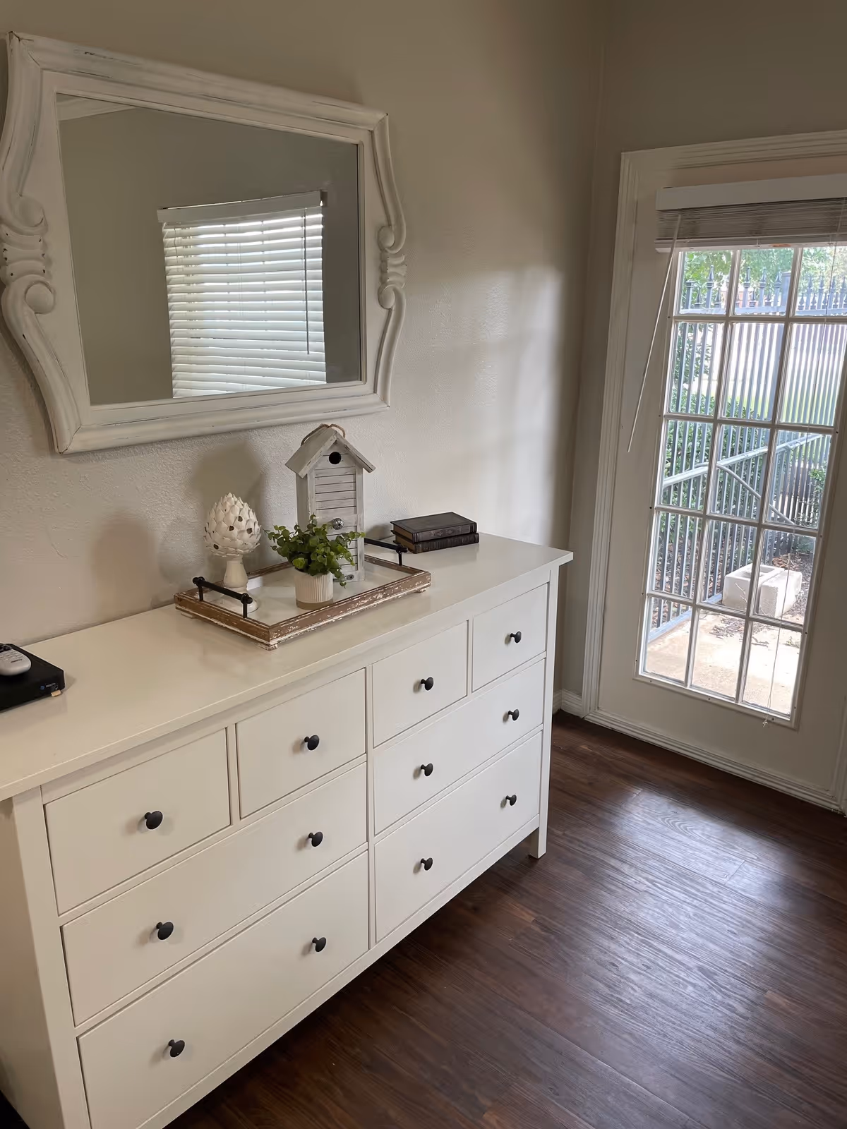 White dresser topped with decorative tray, mirror and small decor next to a glass-paned door showing an outdoor area.