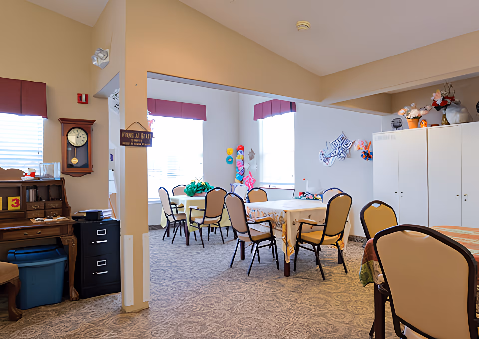A bright room with multiple tables and chairs arranged for group seating. The walls are decorated with colorful crafts and flowers. There is a wooden desk with a clock on the wall and a filing cabinet nearby. The room has large windows with maroon valances letting in natural light.