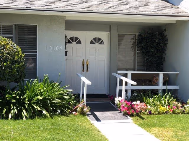 Front entrance of a residential building with white double doors, a small ramp with handrails leading up to the door, surrounded by green plants and colorful flowers. The building number 10109 is visible on the left side of the door.