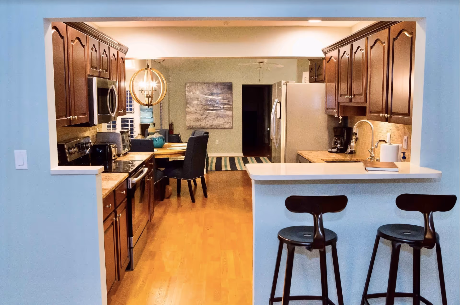 Interior view of a kitchen with wooden cabinets, stainless steel appliances, and a white countertop bar with two black bar stools. Beyond the kitchen is a dining area with a round wooden table, four dark upholstered chairs, a modern chandelier, and a large abstract painting on the wall.