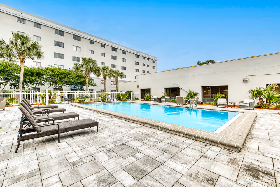 Outdoor swimming pool area with several lounge chairs on a tiled deck, surrounded by a white fence. Palm trees and other greenery are visible along the fence line. A multi-story building and a single-story building with windows and outdoor seating are in the background under a clear blue sky.