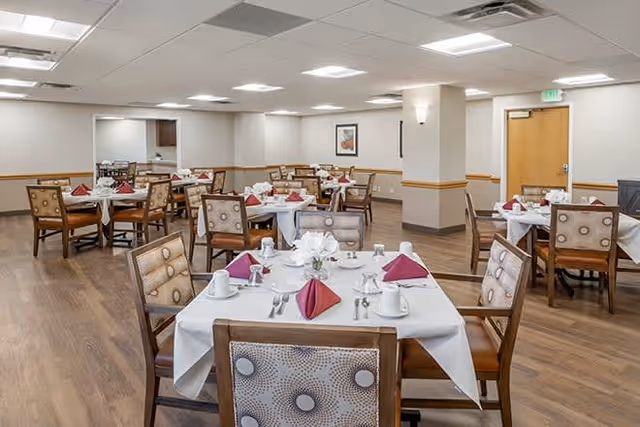A spacious dining room with multiple tables set for a meal. Each table is covered with a white tablecloth and has neatly folded red napkins, white cups, plates, and silverware. The room has wooden flooring, beige walls with a wooden trim, and ceiling lights providing bright illumination. There are several chairs around each table, and a framed picture is visible on one wall.