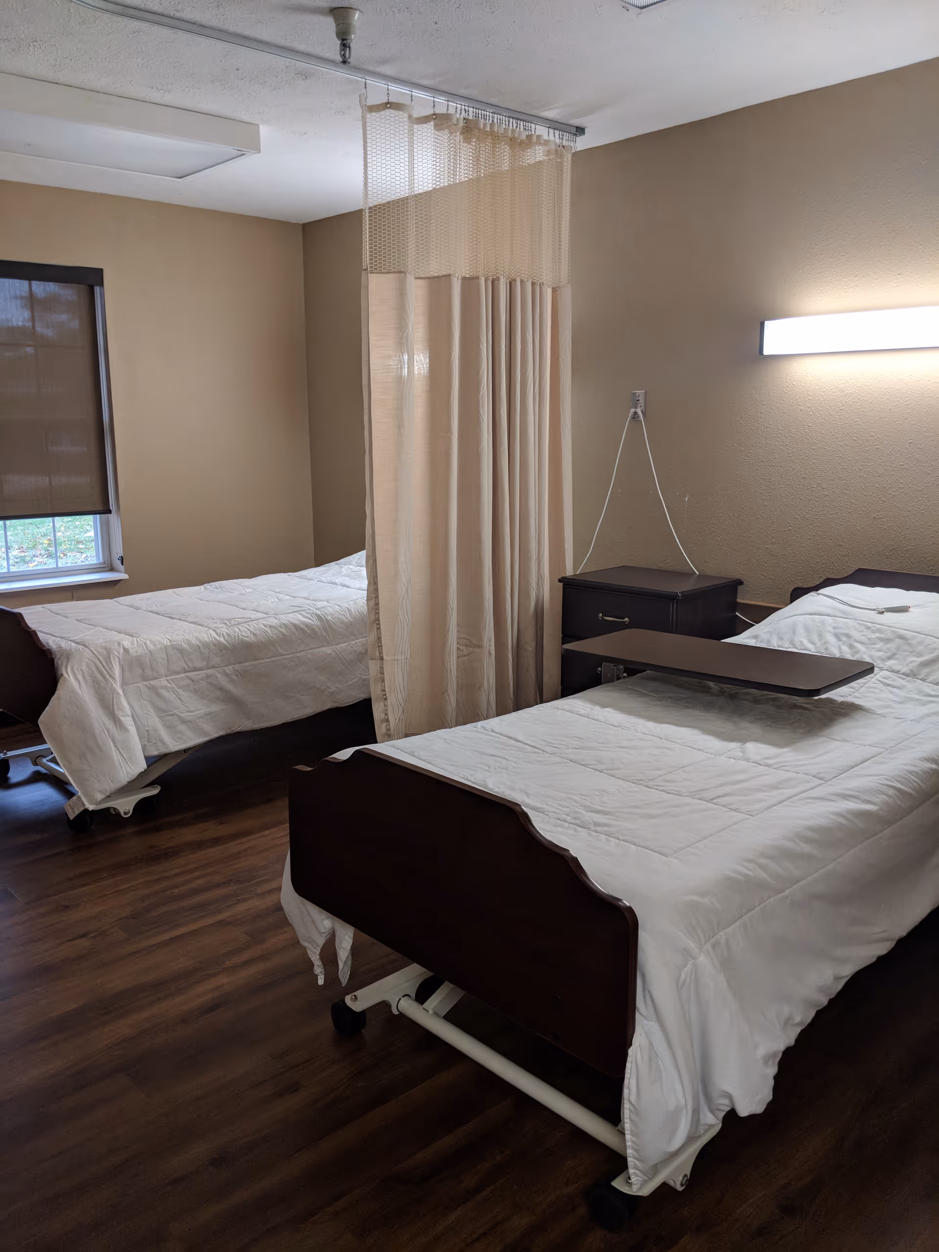 A room with two hospital-style beds separated by a privacy curtain. Each bed has white bedding and a dark wooden headboard and footboard. There is a small bedside table between the beds and a window with a brown shade on the left wall. The floor is wooden, and a wall-mounted light fixture is above the bed on the right.
