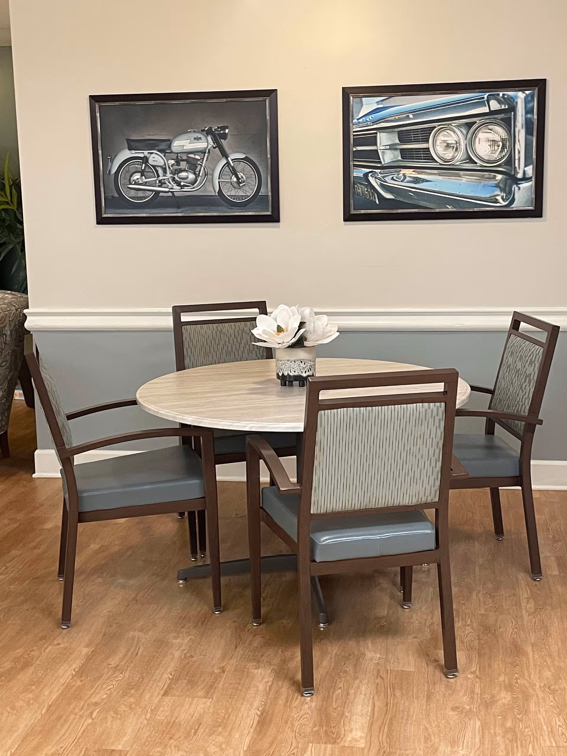 A round wooden table with four chairs around it, each chair having a wooden frame and cushioned seat and backrest. On the table is a small vase with white flowers. The room has light-colored walls with a white chair rail and two framed pictures hanging above the table, one of a motorcycle and the other of a close-up of a car's front grille and headlights. The floor is wooden.