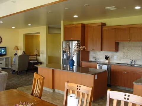 Open-plan community kitchen and dining area with a central island, wooden cabinets, dining table and seating, and a living area visible in the background.