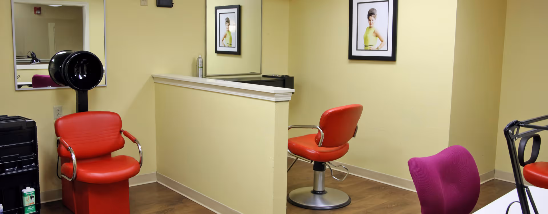Interior view of a salon area with two red salon chairs, a black hair dryer, a purple chair, a large mirror, and a framed picture on a yellow wall.