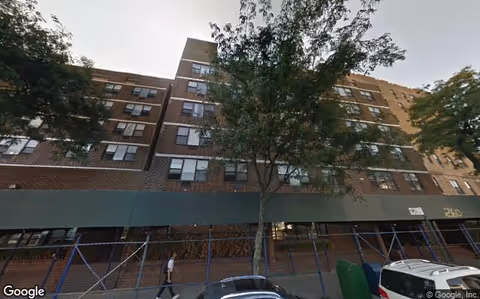 Exterior view of a multi-story brick building with several windows, a tree in front, scaffolding along the sidewalk, and a few parked cars. A person is walking on the sidewalk.