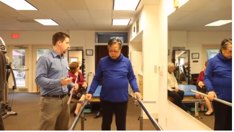 An elderly woman in a blue sweater is using parallel bars for walking exercise while a man in a blue shirt and khaki pants stands beside her holding a clipboard. In the background, a woman in a red jacket is seated on a bench, and an elderly man is also seated. The room appears to be a rehabilitation or therapy area with exercise equipment and large mirrors on the wall.