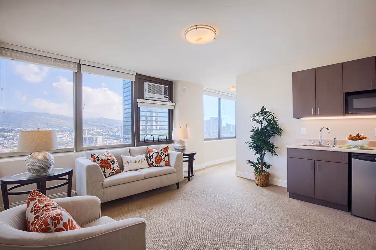 A bright and modern living area with large windows offering a city and mountain view. The room features a beige sofa with floral cushions, a matching armchair, two side tables with lamps, a potted plant, and a kitchenette with dark cabinets, a sink, microwave, and mini fridge.