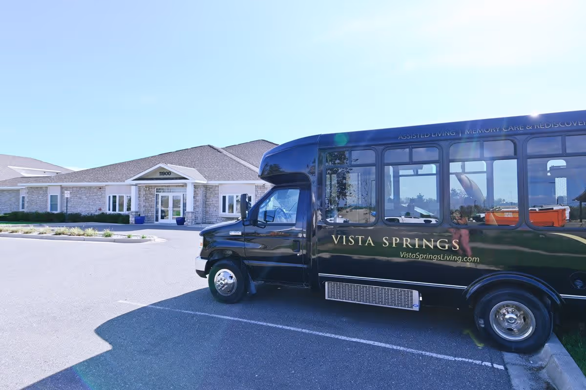 A black Vista Springs shuttle bus parked in front of a single-story stone building with a peaked roof and a covered entrance. The building has the address number 11900 above the entrance. The sky is clear and blue.