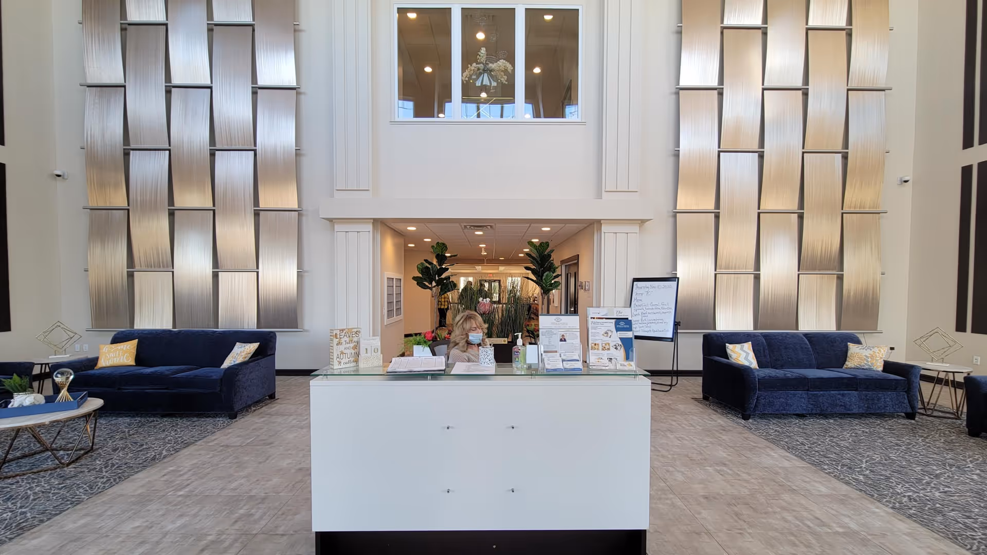 A spacious and modern reception area in a senior living facility with a white front desk where a woman wearing a face mask is seated. Behind the desk are tall decorative wall panels and a large window overlooking an upper floor. On either side of the desk are dark blue sofas with patterned pillows and small side tables. The floor is a combination of carpet and tile, and there are plants and informational displays on the desk.