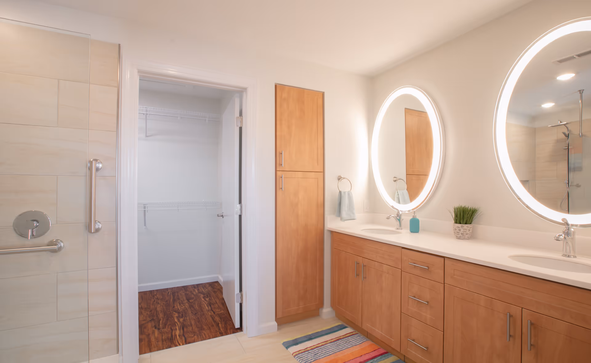 A modern bathroom with a double sink vanity featuring wooden cabinets and two round illuminated mirrors above. To the left is a glass shower door with metal handles and beige tiles. An open door reveals a walk-in closet with wooden flooring and white wire shelving. A colorful striped rug is placed on the light tile floor in front of the vanity.
