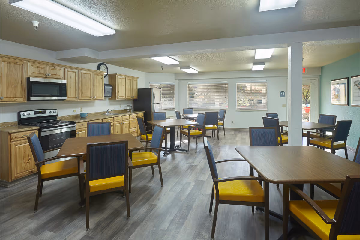A bright dining room with several wooden tables and chairs featuring yellow cushions. The room has a kitchen area with wooden cabinets, a stove, microwave, and refrigerator. Large windows with blinds allow natural light to fill the space, and the floor is covered with wood-like vinyl flooring.