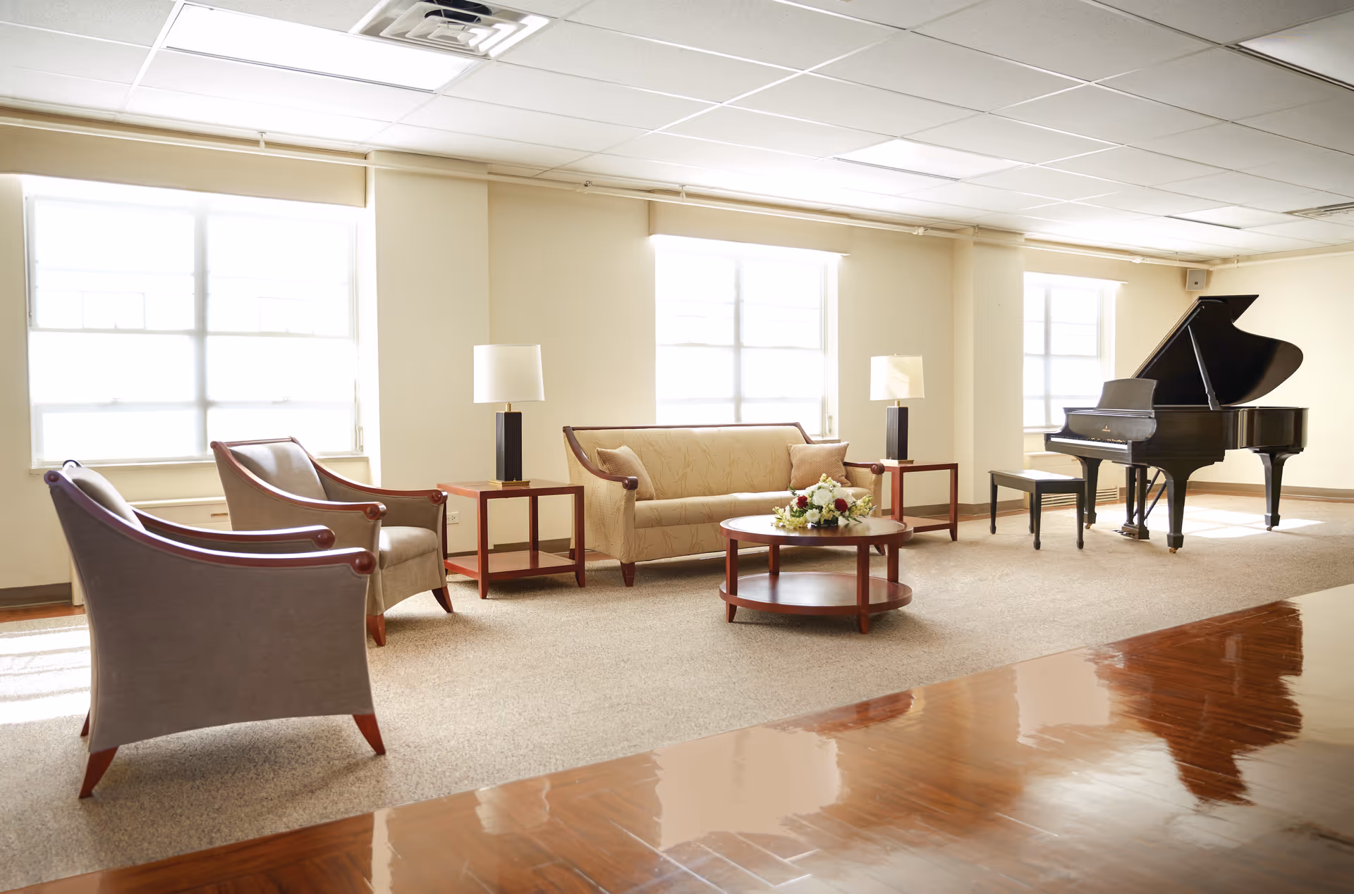 A bright and spacious living room area with large windows letting in natural light. The room features a beige sofa with two cushions, two matching armchairs, two side tables with lamps, a round coffee table with a floral arrangement, and a black grand piano with a bench. The floor is carpeted except for a polished wooden section in the foreground.