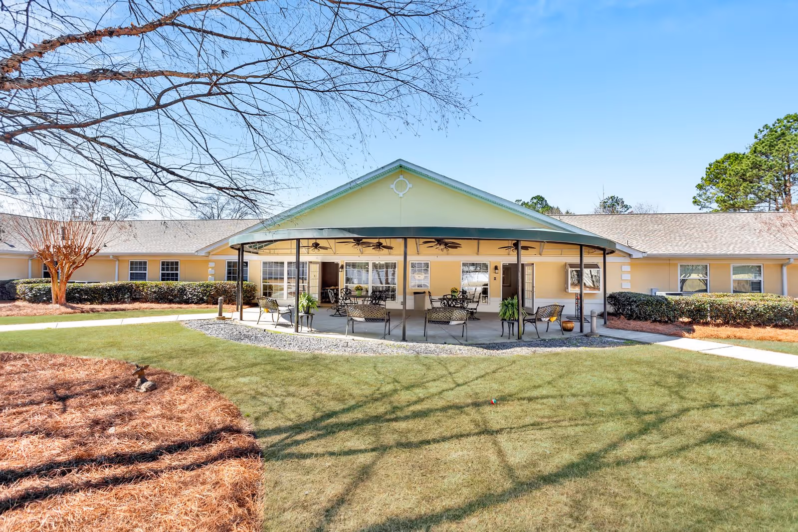 Outdoor patio area of a single-story assisted living facility with a covered seating area featuring metal chairs and tables, surrounded by a well-maintained lawn and landscaped bushes under a clear blue sky.