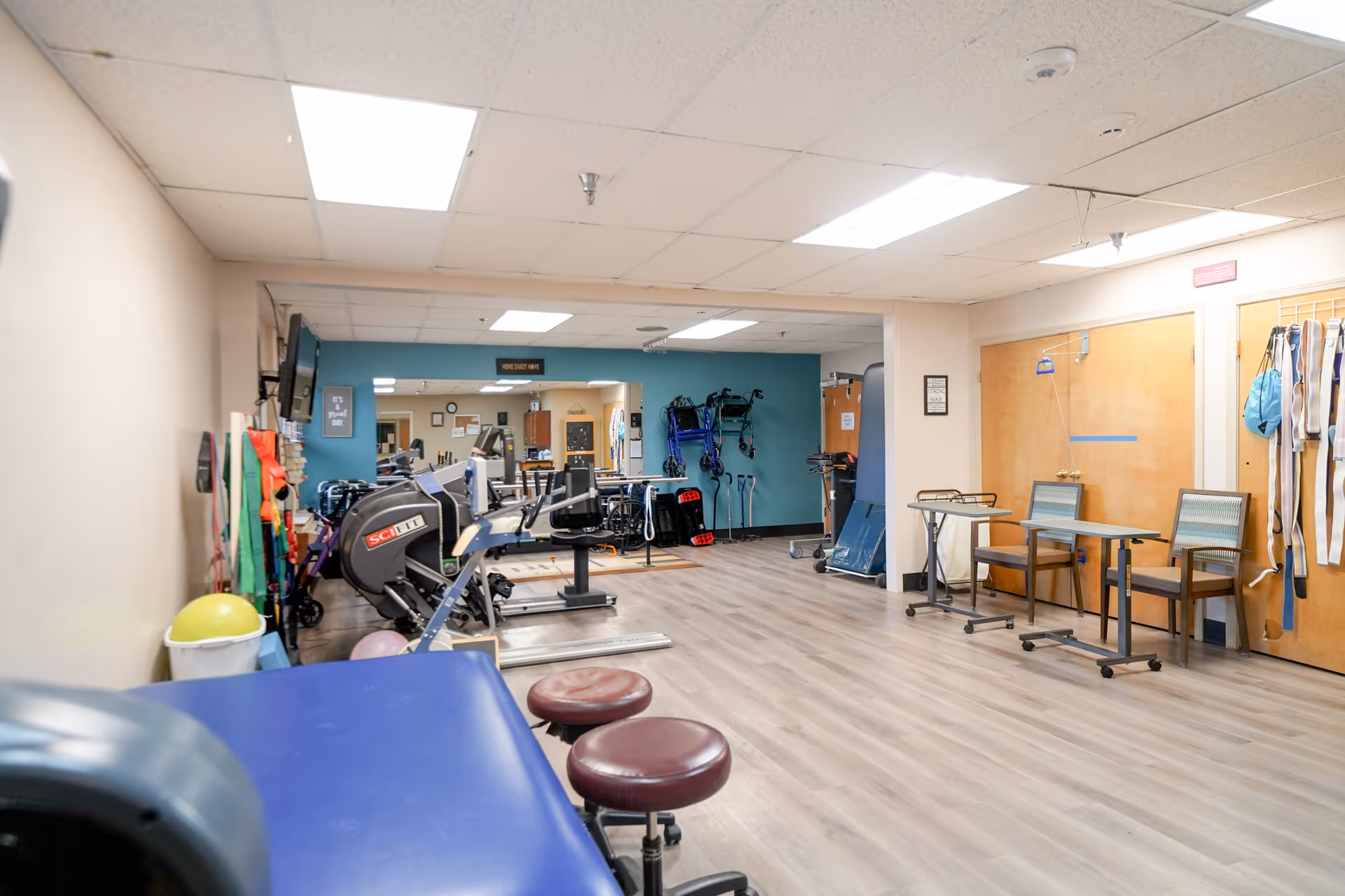Interior view of a rehabilitation or physical therapy room with exercise equipment, therapy tables, chairs, and various mobility aids such as walkers and canes. The room has light-colored walls, wood flooring, and fluorescent ceiling lights.