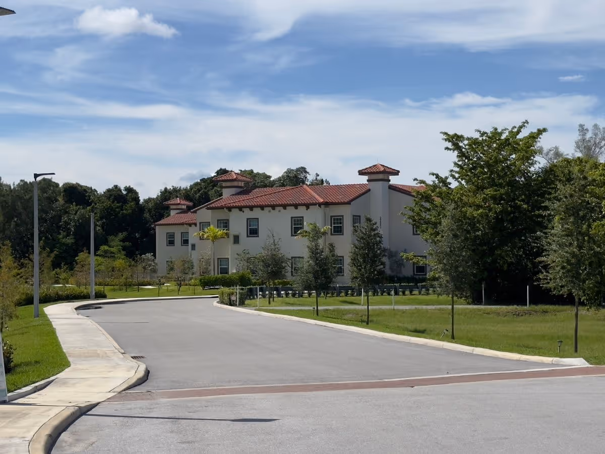 Two-story Mediterranean-style building with red tile roof at the end of a curved driveway, surrounded by trees and lawn under a blue sky.