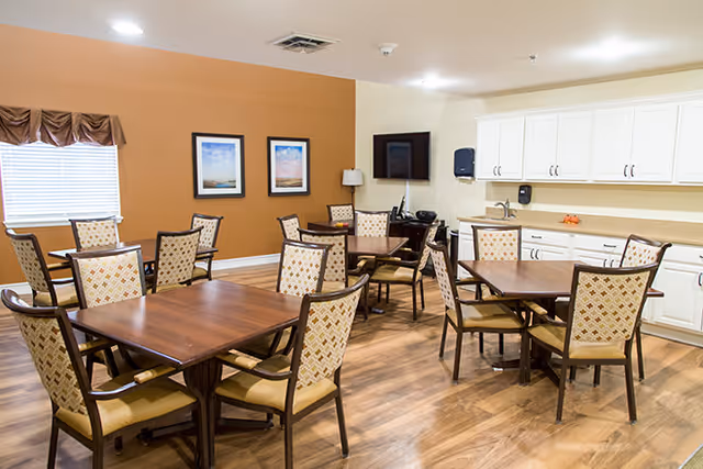 A bright communal dining room with several wooden tables and upholstered chairs, a kitchenette with white cabinets, and a wall-mounted TV.