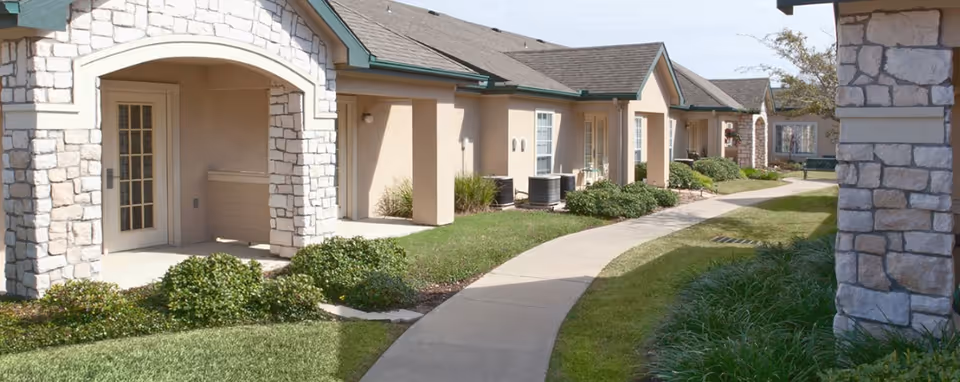 A curved concrete walkway runs alongside a single-story building with beige walls and stone accents. The building has multiple doors and windows, with green shrubbery and grass lining the path. The sky is clear and blue.