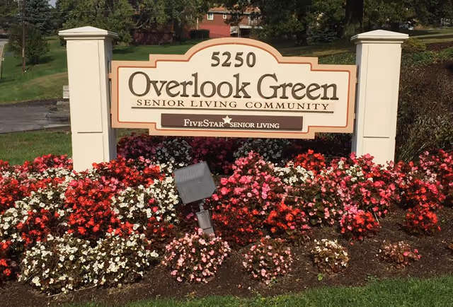 Entrance sign reading "5250 Overlook Green Senior Living Community" surrounded by red, pink, and white flower beds.