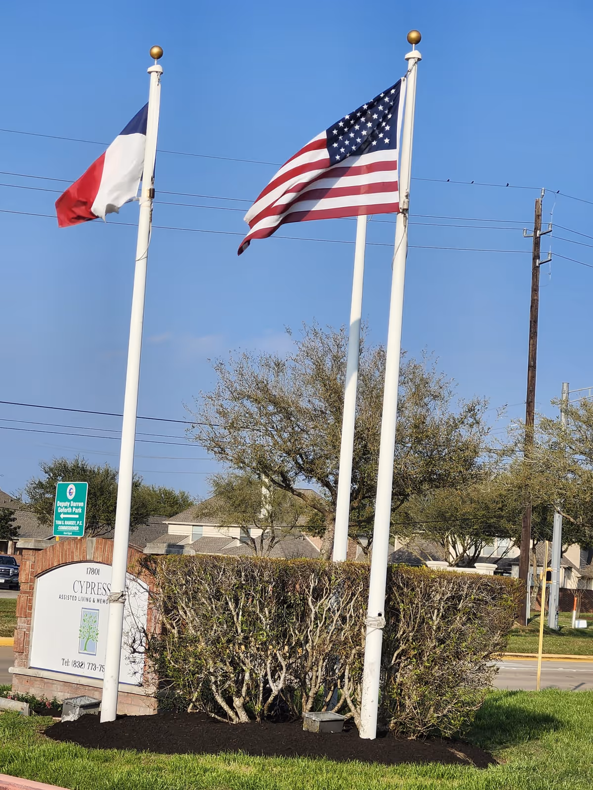 Two flagpoles with the Texas state flag and the United States flag flying against a clear blue sky, with a sign below reading Cypress Assisted Living & Memory Care, surrounded by trimmed bushes and a grassy area.