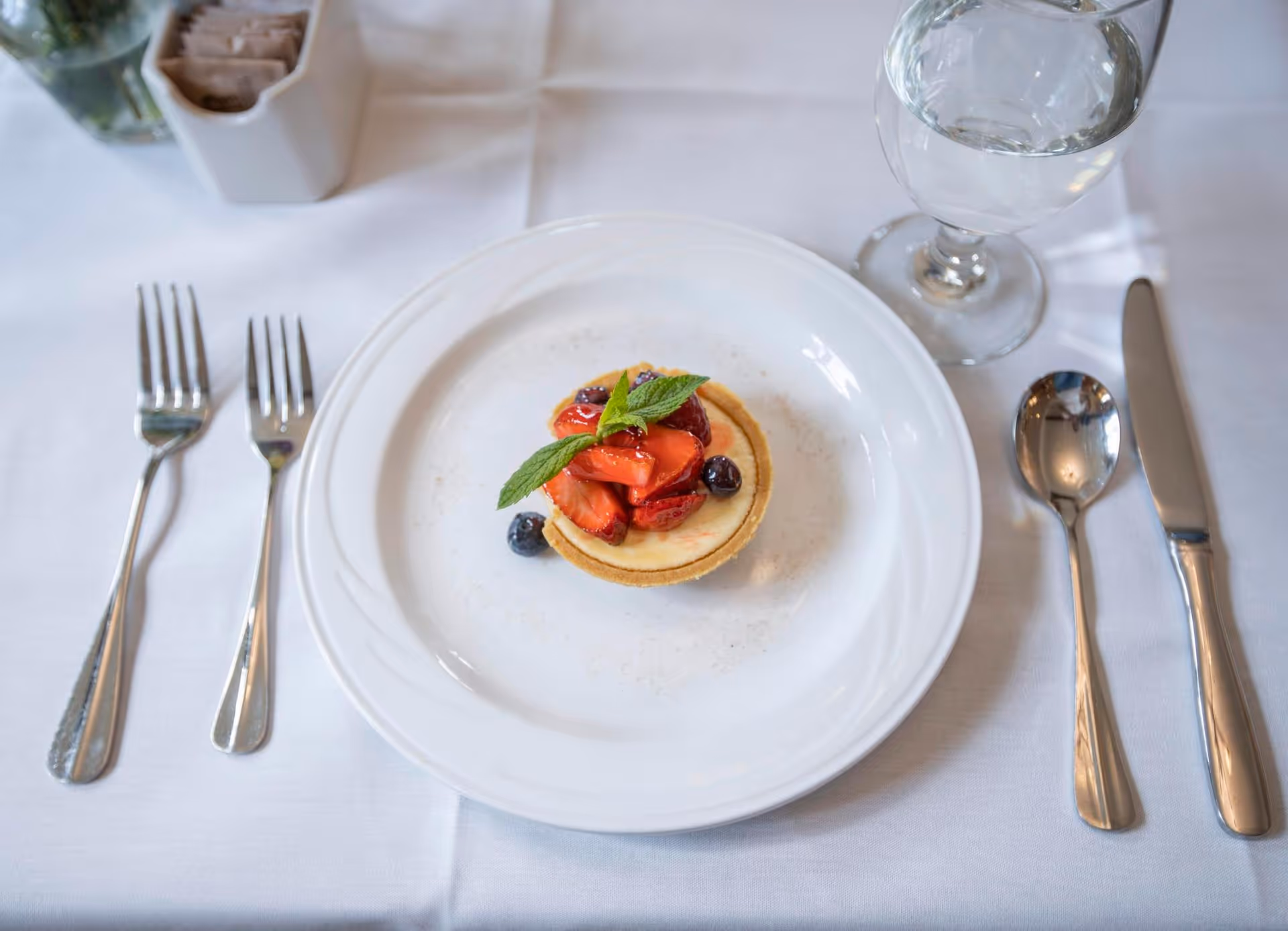 A small fruit tart topped with strawberries, blueberries, and a mint leaf on a white plate, set on a white tablecloth with two forks on the left and a knife, spoon, and glass of water on the right.