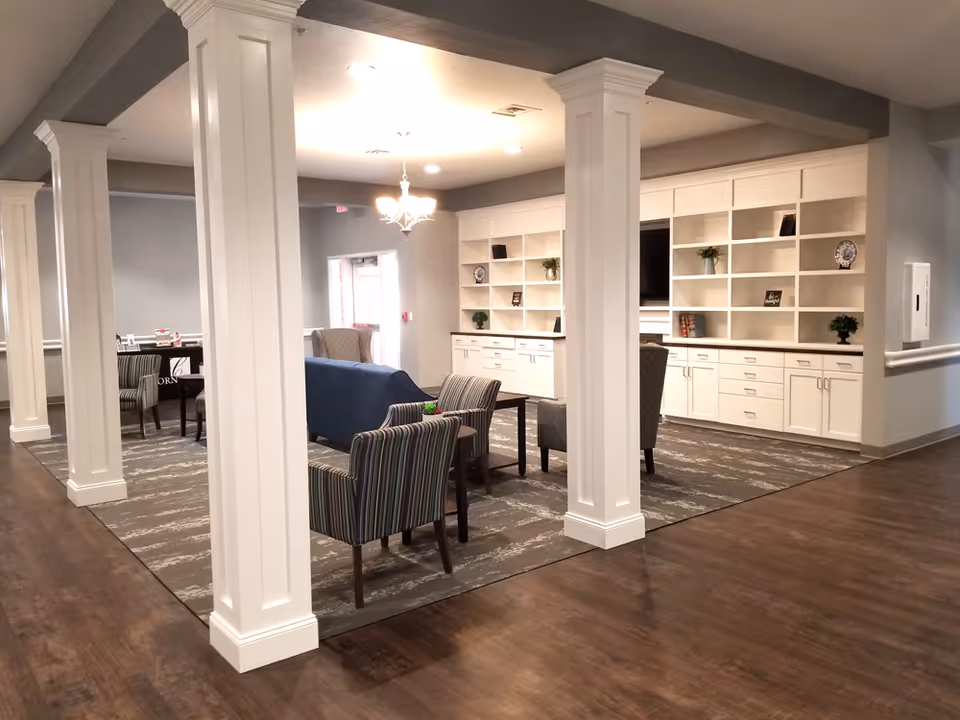 A spacious and well-lit common area in a senior living facility featuring white pillars, a chandelier, built-in white shelves with decorative items, a flat-screen TV, and various seating options including striped armchairs and a blue sofa on a patterned carpet.