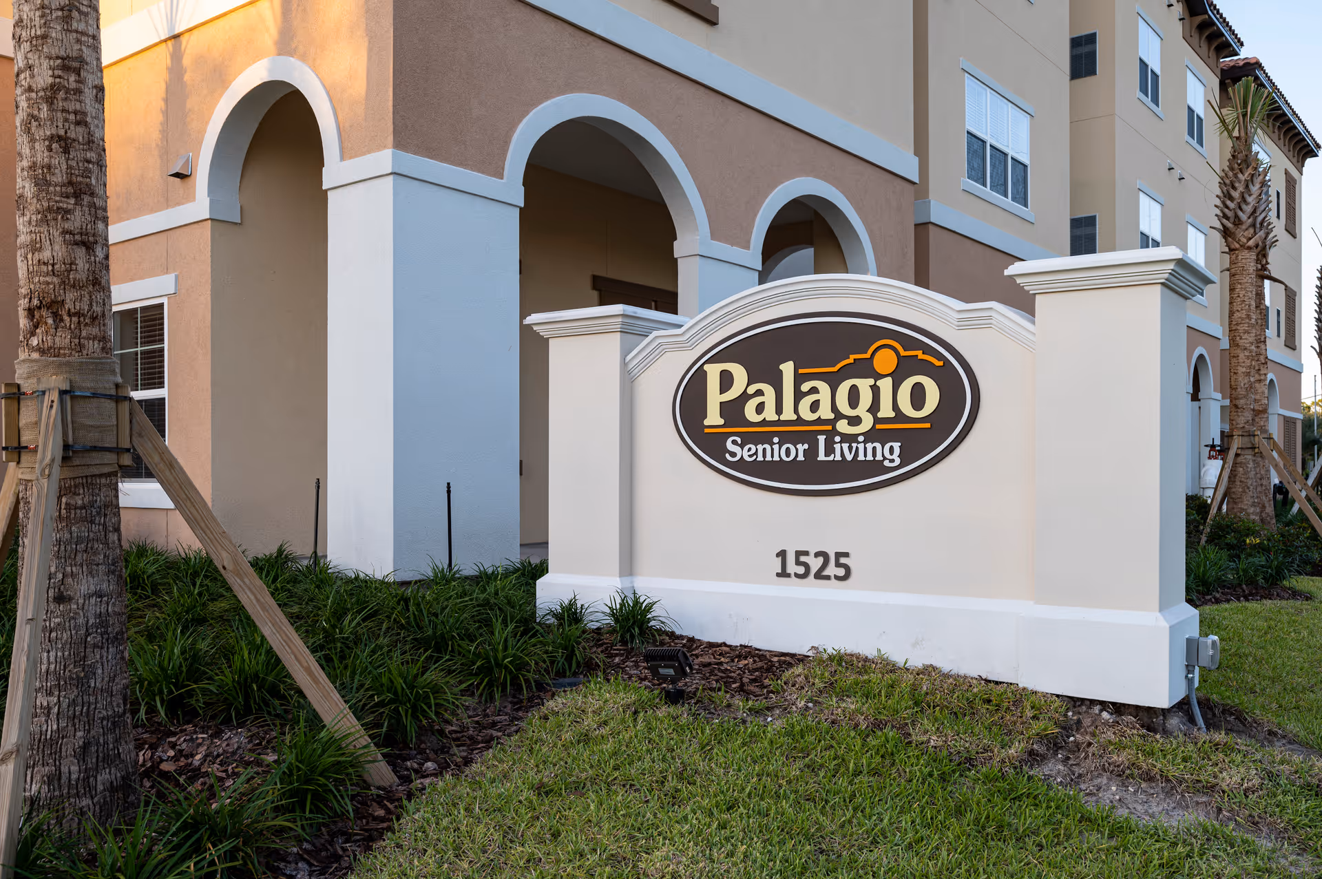 Exterior view of Palagio Senior Living facility showing the building's beige and white facade with arched architectural details, palm trees, green landscaping, and a prominent sign displaying the facility name and address number 1525.