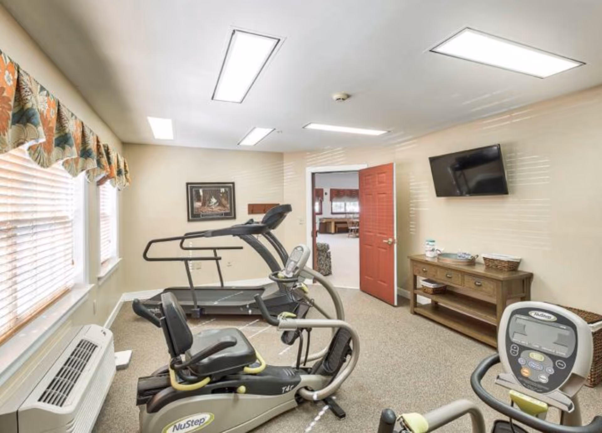 A small fitness room with exercise equipment including a treadmill and two recumbent bikes. The room has beige walls, carpeted floor, a wall-mounted TV, a wooden console table with baskets, and windows with floral valances letting in natural light. An open red door leads to another room.